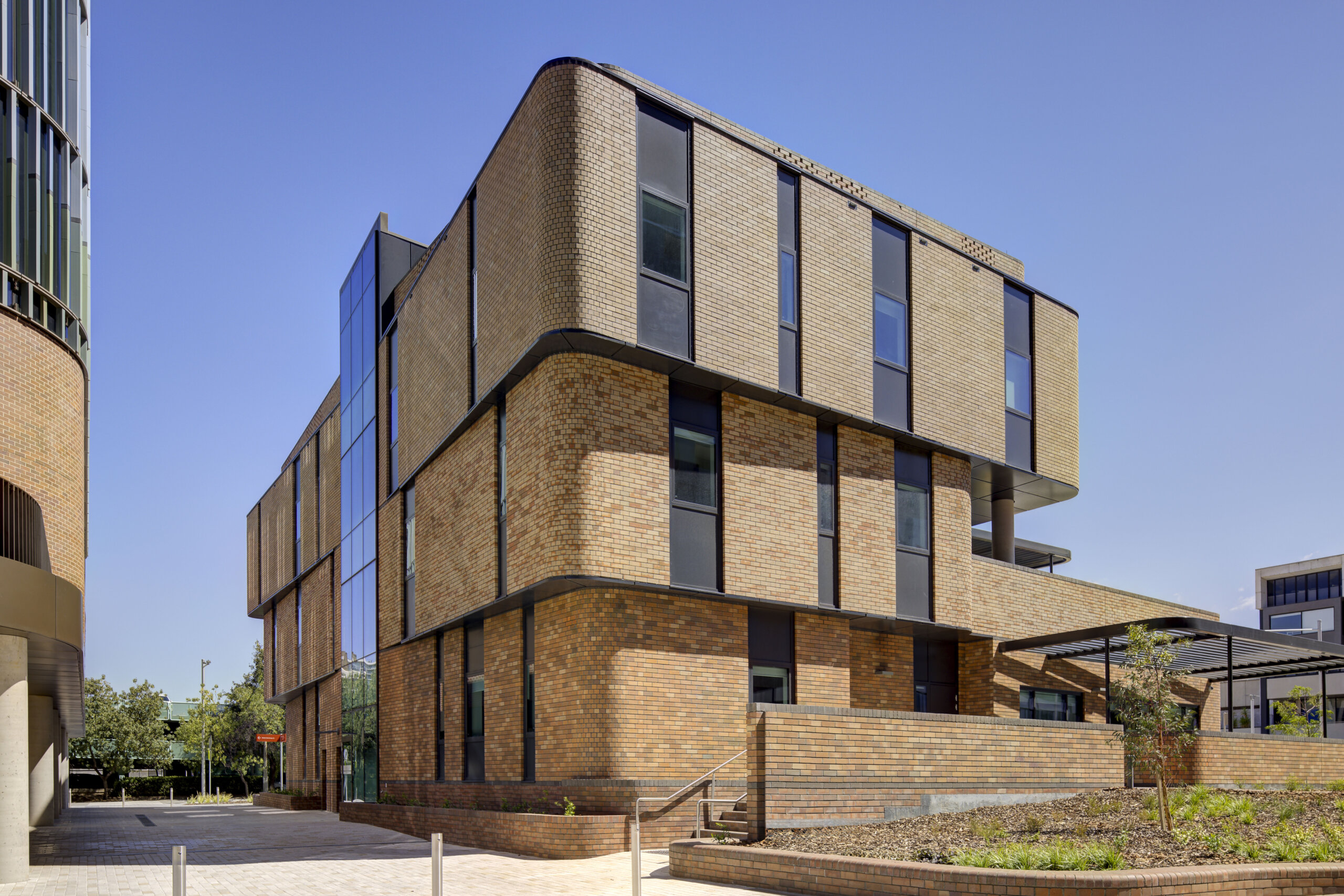 A modern three-storey building constructed of tan brick with dark-framed windows and a glass-panelled corner section under a clear blue sky.