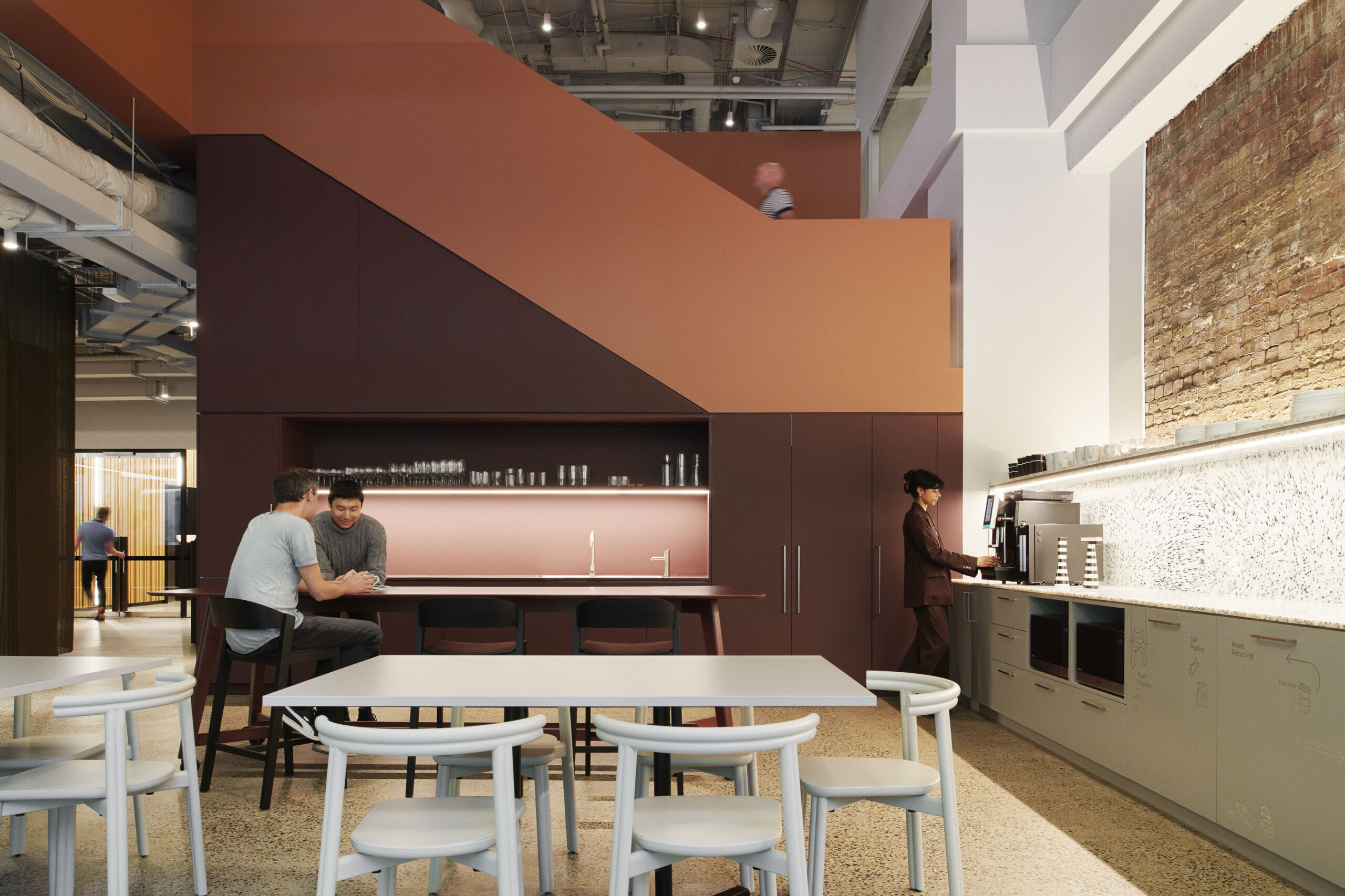 Office break room with a central kitchen island, seating area, and a large orange staircase leading to an upper level, showcasing Gray Puksand Melbourne Design Week aesthetics.