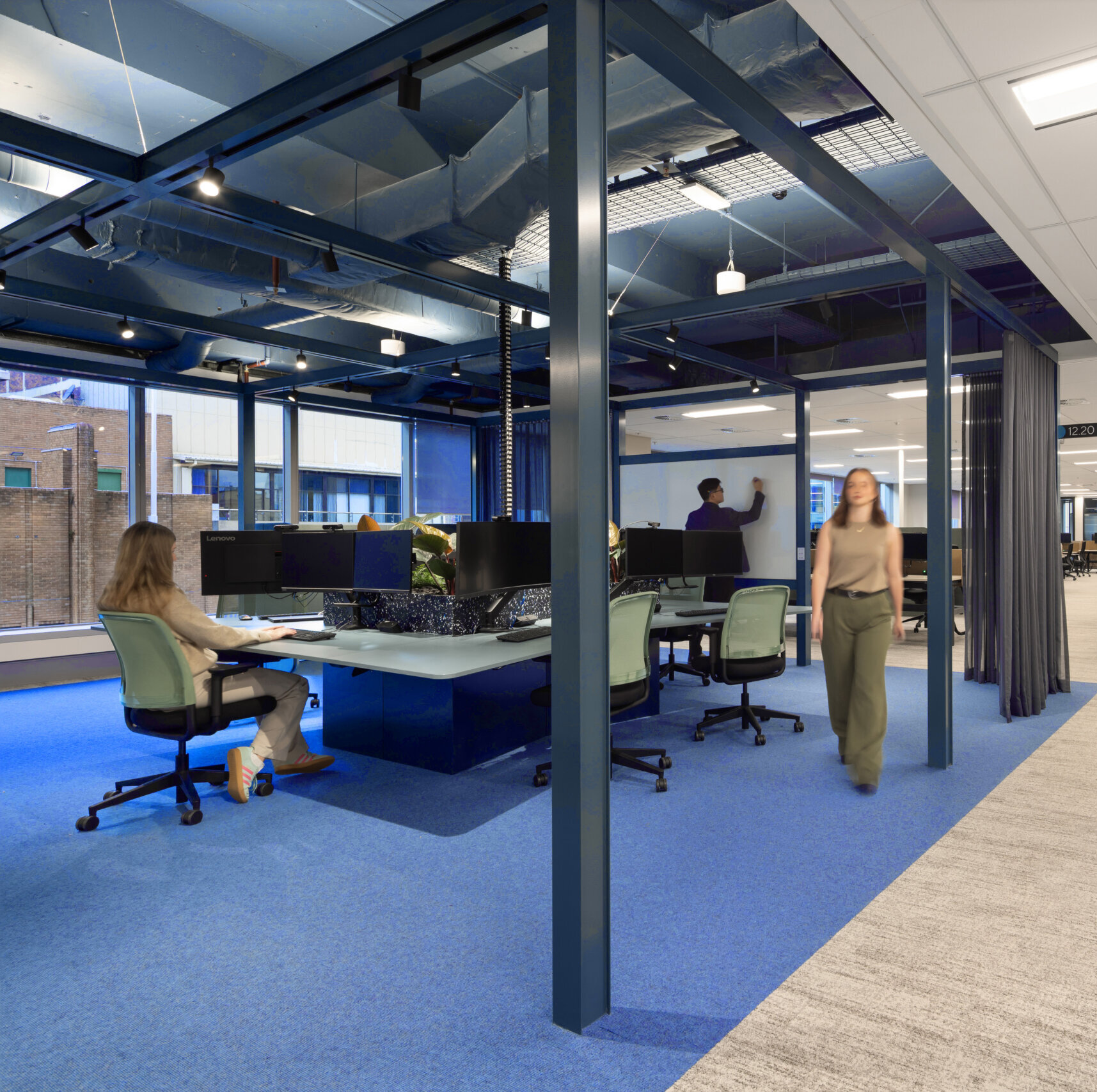 Employees work in a modern office space featuring blue and grey Puksand carpeting, exposed ceiling beams, and a mix of open workstations and semi-private areas designed for Melbourne Design Week.
