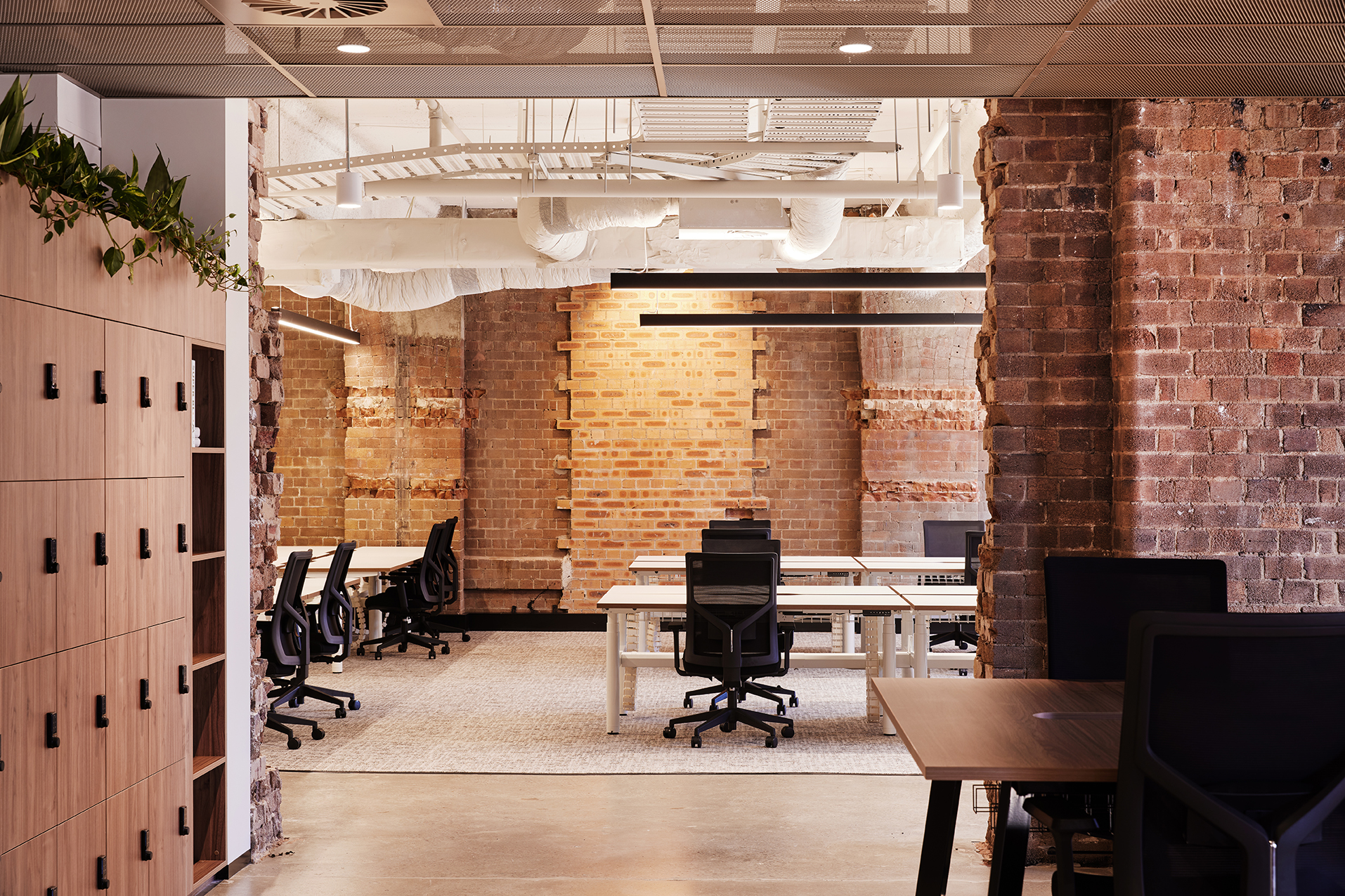 An open-plan office featuring Melbourne design week aesthetic, with exposed brick walls, rows of desks with rolling black chairs, and light wood storage lockers on the left.